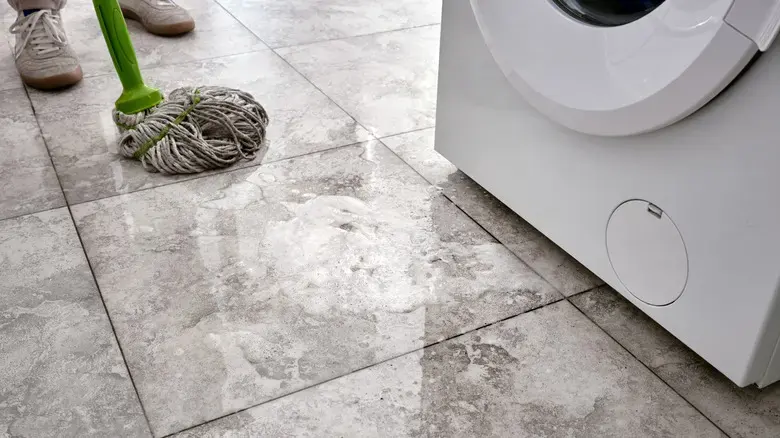 Mop and bucket on a wet tile floor beside a front-load washing machine after a leak