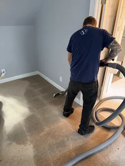 Technician running an extraction wand and hose over wet carpet in an emptied room near Garner, NC